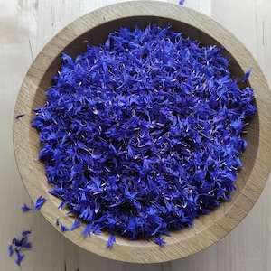 May include: A round, light-coloured wooden bowl filled with a large quantity of dried, vibrant blue cornflower petals. The petals are small, thin, and have a deep, saturated blue colour, with a few loose petals scattered outside the bowl.