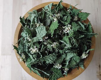 Può includere: Una ciotola di legno piena di foglie verdi essiccate e piccoli fiori bianchi. Le foglie hanno un aspetto strutturato e la ciotola poggia su una superficie di legno chiaro. La disposizione suggerisce un tema naturale ed erboristico.