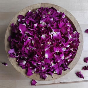 May include: A round wooden bowl filled with dried rose petals in deep shades of magenta and purple. The petals have subtle white and cream-coloured edges. Several loose petals are scattered around the bowl on the light-coloured surface.