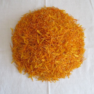 Pu&ograve; includere: Un mucchio di petali di calendula arancioni secchi su una superficie bianca.