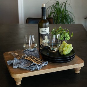 Puede incluir: Una bandeja de servir de madera con un acabado marrón claro y cuatro patas. La bandeja contiene dos copas de vino, una botella de vino 19 Crimes, una pequeña planta en maceta, un racimo de uvas verdes y dos platos negros.