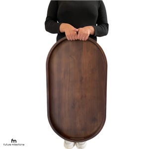 Puede incluir: Una gran bandeja ovalada de madera con acabado marrón oscuro. La bandeja tiene un borde elevado y la sostiene una persona. La bandeja mide aproximadamente 76 cm de largo y 46 cm de ancho.