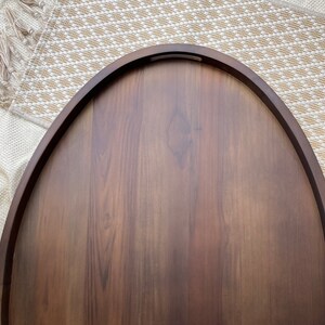 Puede incluir: Una bandeja de madera ovalada de color marr&oacute;n oscuro, con un borde curvo y un hueco para el asa. La bandeja tiene una superficie lisa y pulida y est&aacute; colocada sobre una alfombra beige tejida con flecos.