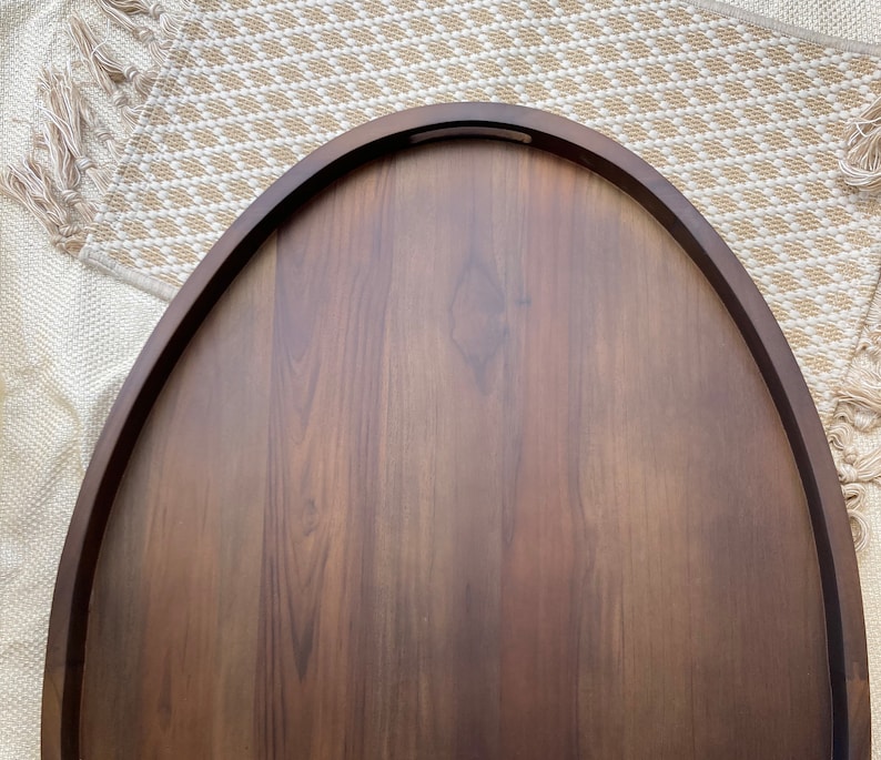 Puede incluir: Una bandeja de madera ovalada de color marr&oacute;n oscuro, con un borde curvo y un hueco para el asa. La bandeja tiene una superficie lisa y pulida y est&aacute; colocada sobre una alfombra beige tejida con flecos.