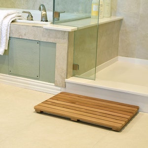 Könnte beinhalten: Eine hellbraune Holz-Badematte mit Lamellenmuster. Die Matte liegt auf einem weißen Fliesenboden in einem Badezimmer.