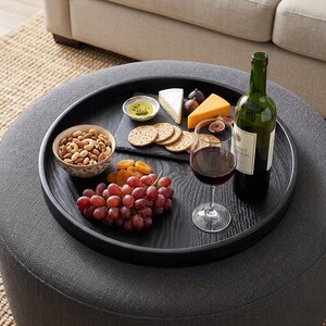 Può includere: Un vassoio di legno nero con una selezione di snack e bevande. Presenta una ciotola di noci, uva, formaggio, cracker, fichi e una bottiglia di vino. Un bicchiere di vino rosso è posto accanto alla bottiglia.