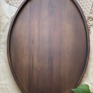 Puede incluir: Bandeja ovalada de madera para servir con acabado marr&oacute;n oscuro. La bandeja tiene un borde elevado y asas integradas. La veta de la madera es visible, lo que a&ntilde;ade textura a la superficie. La bandeja est&aacute; dise&ntilde;ada para servir alimentos o exhibir art&iacute;culos.