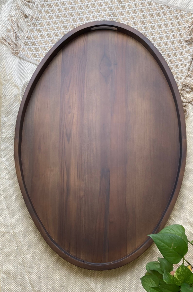 Puede incluir: Bandeja ovalada de madera para servir con acabado marr&oacute;n oscuro. La bandeja tiene un borde elevado y asas integradas. La veta de la madera es visible, lo que a&ntilde;ade textura a la superficie. La bandeja est&aacute; dise&ntilde;ada para servir alimentos o exhibir art&iacute;culos.