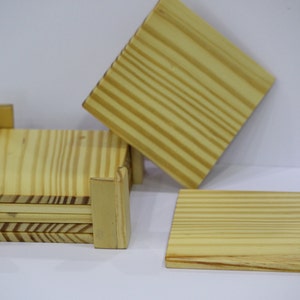 Puede incluir: Conjunto de seis posavasos de madera en acabado de madera natural. Los posavasos son cuadrados y tienen un borde elevado. Los posavasos están apilados en un soporte, con un posavasos apoyado en la pila.