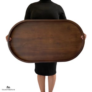 May include: A large, oval-shaped wooden serving tray with a dark brown finish. The tray is being held by a person, but the person's hands are not visible in the image.