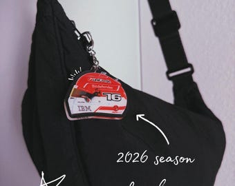 Könnte beinhalten: Eine schwarze Umhängetasche mit einem angehängten Schlüsselanhänger in Form eines rot-weißen Rennhelms. Die Grafik des Helms zeigt die Nummer 16 sowie den Text UniCredit, Bitdefender und IBM. Das Bild enthält zudem den handschriftlichen Text 2026 season keychain und drei gezeichnete Sterne.