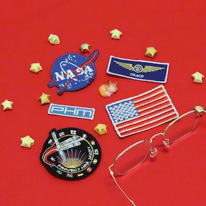 Puede incluir: Una colección de parches bordados que incluye un logotipo azul de la NASA, un parche rectangular azul de Grace con alas doradas, un parche pequeño de PHM, un parche rectangular de la bandera estadounidense y un parche circular de Project Hail Mary.