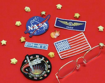 Puede incluir: Una colección de parches bordados que incluye un logotipo azul de la NASA, un parche rectangular azul de Grace con alas doradas, un parche pequeño de PHM, un parche rectangular de la bandera estadounidense y un parche circular de Project Hail Mary.