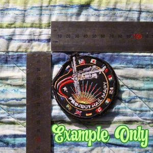 May include: A circular embroidered patch featuring a space station graphic, a sunburst, and the text "PROJECT HAIL MARY" with small international flags along the border. A metal ruler is visible, showing the patch is approximately 7.5 cm wide.