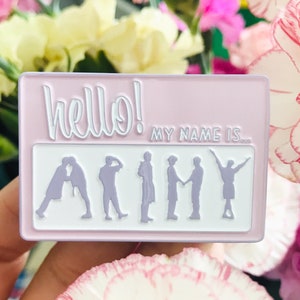 Puede incluir: Un pin de esmalte rosa con el texto "hello! my name is... ARMY" en blanco. Siete siluetas de personas moradas están dispuestas en una fila debajo del texto.