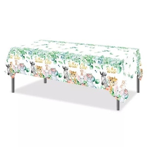 May include: White plastic tablecloth with a jungle animal theme. The tablecloth features a green leafy pattern with a variety of animals including a zebra, lion, elephant, and giraffe. The text "Wild One" is repeated throughout the tablecloth.