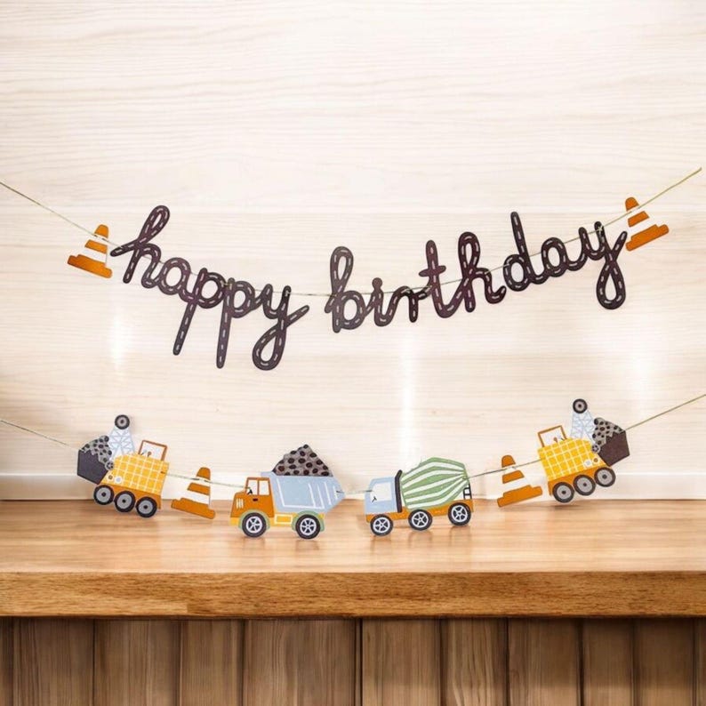 以下が含まれることがあります： 「happy birthday」の文字がダークブラウンの筆記体で書かれたバースデーバナー。バナーには、トラック、トラフィックコーン、装飾要素などの建設をテーマにした切り抜きが含まれており、すべて細い紐で繋がれています。