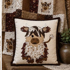 Pode incluir: Uma almofada quadrada com uma aplicação de tecido de uma vaca Highland em tons de castanho, creme e padrões florais sobre um fundo acolchoado creme. A almofada tem uma borda castanha escura e exibe o texto: PERFECT FOR QUILTS, PILLOWS, WALL HANGINGS & HOME DECOR!