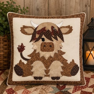 May include: A square throw cushion featuring a central appliqué of a Highland cow in shades of brown, tan, and cream. The cow has a shaggy coat, curved horns, and a tail with a dark brown tip. The cushion has a dark brown patterned border.