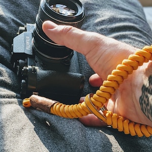 Może przedstawiać: Czarny aparat fotograficzny z żółtym paskiem na aparat. Aparat ma osłonę obiektywu, a pasek jest wykonany z plecionej liny.