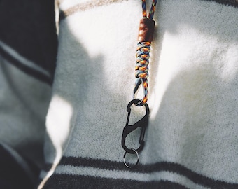 Op de afbeelding: Een keycord met een veelkleurig gevlochten koord en een bruin lederen accent, verbonden aan een zwarte metalen karabijnhaak en een kleine zilveren sleutelring, hangend tegen een wit en donkergrijs gestreepte gebreide trui.
