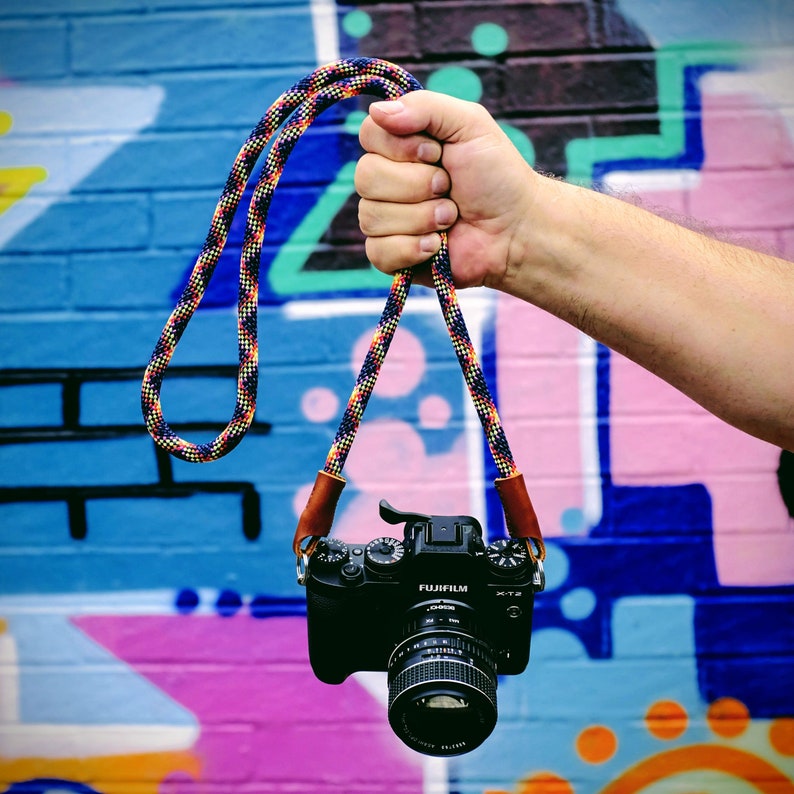 Op de afbeelding: Een zwarte camera met een kleurrijke cameraband die aan een hand hangt tegen een kleurrijke graffitimuur. De camera heeft de tekst "FUJIFILM" en "XT2" erop.
