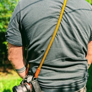 Op de afbeelding: Een persoon draagt een grijs T-shirt met een gele en bruine cameraband. Een zwarte camera met een lens is aan de band bevestigd.