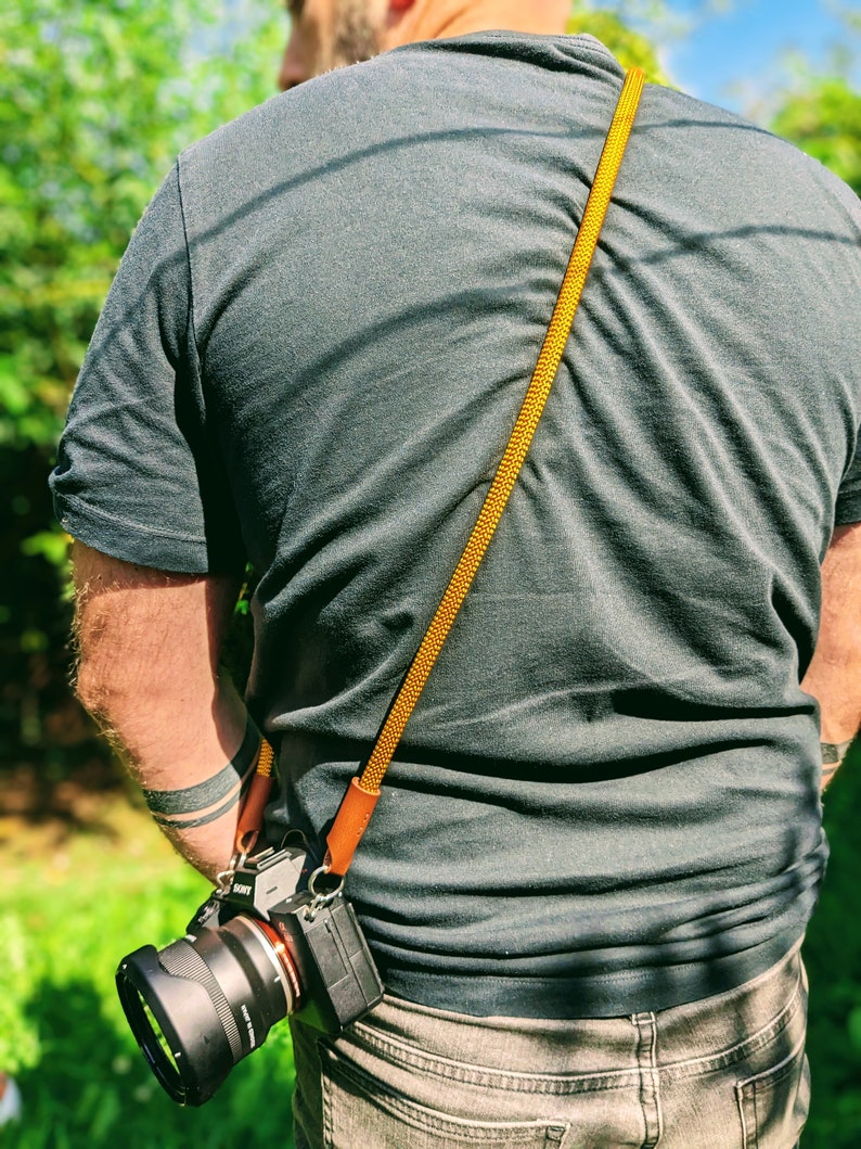 Op de afbeelding: Een persoon draagt een grijs T-shirt met een gele en bruine cameraband. Een zwarte camera met een lens is aan de band bevestigd.