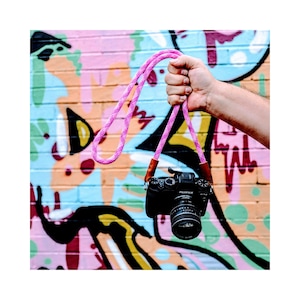 Op de afbeelding: Een roze cameraband met bruine leren accenten die aan een hand hangt tegen een kleurrijke graffitimuur.