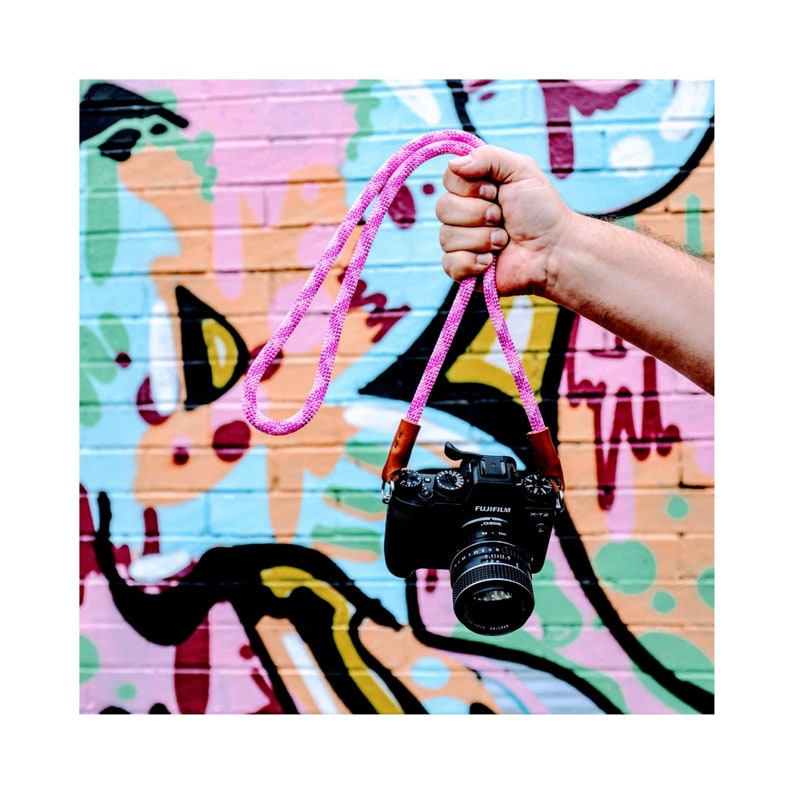 Op de afbeelding: Een roze cameraband met bruine leren accenten die aan een hand hangt tegen een kleurrijke graffitimuur.