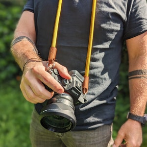 Op de afbeelding: Een persoon draagt een zwart T-shirt en een blauwe jeans en houdt een zwarte Sony-camera vast met een gele en bruine cameraband. De camera heeft een zwarte lens met de tekst "SONY" en "DIM. 050 mm 1:2.8" erop gedrukt.