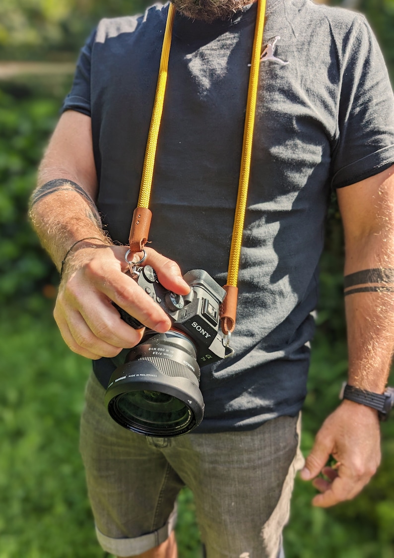 Op de afbeelding: Een persoon draagt een zwart T-shirt en een blauwe jeans en houdt een zwarte Sony-camera vast met een gele en bruine cameraband. De camera heeft een zwarte lens met de tekst "SONY" en "DIM. 050 mm 1:2.8" erop gedrukt.