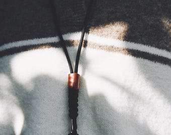 Op de afbeelding: Een zwart koord met een gladde bruine cilindrische kraal, een gevlochten zwart gedeelte en een zwarte metalen karabijnhaak met een kleine ronde ring aan de onderkant, gedragen over een bruin en wit horizontaal gestreepte trui.