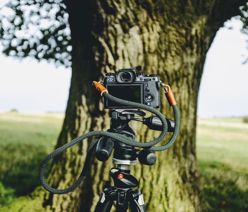Op de afbeelding: Een zwarte camera met een lens en een band is gemonteerd op een statief met een groene band. De camera is geplaatst voor een grote boomstam.
