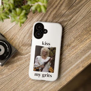 Könnte beinhalten: Eine weiße Smartphone-Hülle mit einer rechteckigen Grafik einer Person mit blonden Haaren, die eine weiße Rüschenbluse trägt und ein langes Gewehr hält. Der Text "kiss" steht über dem Bild und "my grits" steht unter dem Bild.