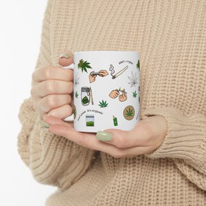May include: White ceramic mug with a green, black, and brown design featuring various cannabis-related illustrations, including leaves, joints, and a jar of marijuana. The text on the mug reads "100% vegan" and "it's organic".