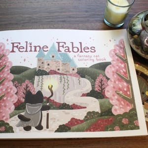 May include: A coloring book titled "Feline Fables" featuring a fantasy cat illustration. The cover art depicts a black cat with a staff, a castle, and a winding path. Pink and green trees frame the scene. A candle and a succulent terrarium are in the background.