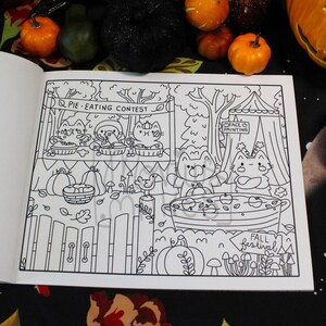 May include: Black and white illustration from a coloring book featuring a fall festival scene. The scene includes a pie-eating contest, face painting, and a large pot with apples. The words "Pie-Eating Contest" and "Fall Festival" are visible.