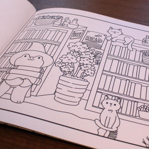May include: A black and white coloring page featuring a frog wearing a wizard hat and holding books, a potted plant, a bookshelf with books and a sign that says "READ", and two cats.
