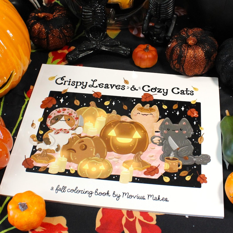 May include: A fall-themed coloring book titled "Crispy Leaves & Cozy Cats" featuring illustrations of pumpkins, cats, and autumn leaves. The cover art includes the text "a fall coloring book by Movius Makes."
