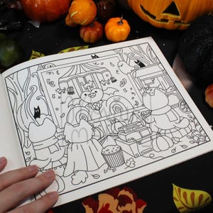 May include: Open coloring book page with a Halloween theme. The black and white illustration features cartoon cats in costumes, a haunted house, pumpkins, and a popcorn stand. The page is surrounded by pumpkins and fall foliage.