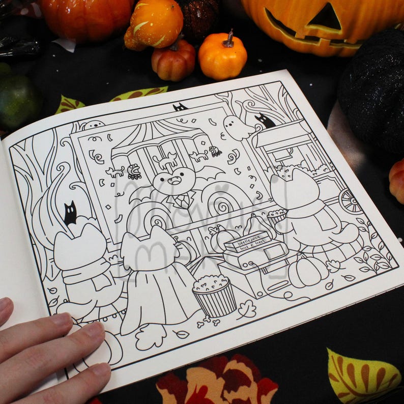 May include: Open coloring book page with a Halloween theme. The black and white illustration features cartoon cats in costumes, a haunted house, pumpkins, and a popcorn stand. The page is surrounded by pumpkins and fall foliage.