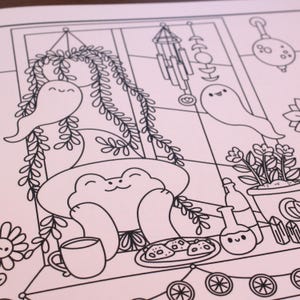 May include: A black and white line drawing of a frog sitting in a window with a plant, a ghost, and a wind chime. The frog is wearing a witch's hat and holding a cup of tea. There are cookies on a plate in front of the frog.