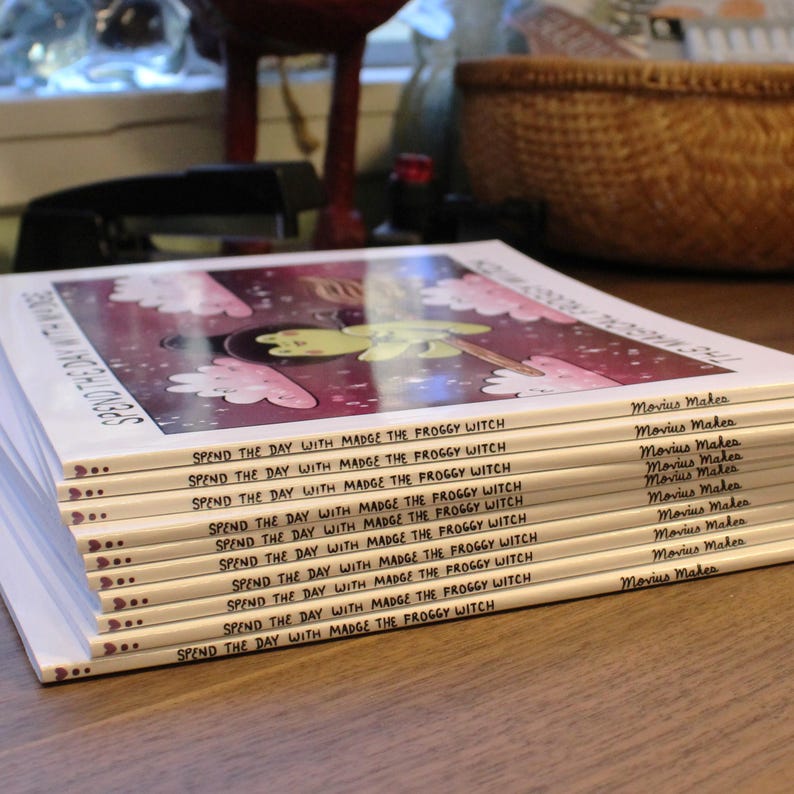 May include: A stack of ten paperback books with a colorful illustration of a frog on a broomstick on the cover. The title of the book is "Spend the Day with Madge the Froggy Witch". The author is Movius Makes.