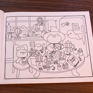 May include: A black and white line drawing of a kitchen scene with a frog, a bear, and a ghost. The frog is sitting at a table with a cup of tea and a plate of food. The bear is sitting in a chair next to the table. The ghost is floating in the air above the table. The table is set with a book that says "Herbal Magic".
