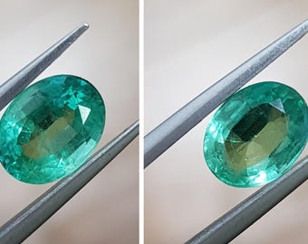 May include: A vibrant green emerald gemstone, oval-shaped, held by tweezers. The emerald has a slightly yellowish hue and is translucent.