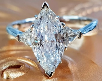 Puede incluir: Anillo de compromiso de plata con un diamante de talla marquesa transparente. El anillo presenta un gran diamante central de talla marquesa, flanqueado por dos diamantes triangulares más pequeños. El diseño es clásico y elegante.