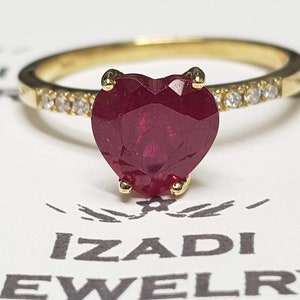 Puede incluir: Un anillo de oro con una piedra preciosa de rubí en forma de corazón y una banda de pequeños diamantes.