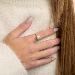 May include: A close-up of a hand wearing a silver ring with a large, clear gemstone. The ring is on the middle finger of the hand.