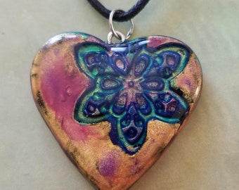 Könnte beinhalten: Eine herzförmige Anhänger-Halskette mit einem schwarzen Band. Der Anhänger ist aus Harz und zeigt ein farbenfrohes Mandala-Design in Blautönen, Grüntönen, Rosa und Gold.
